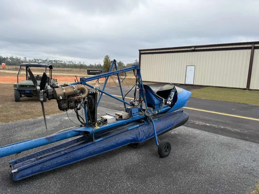 A blue ultralight aircraft on floats, with its engine and propeller visible, parked on an airfield next to a golf cart.