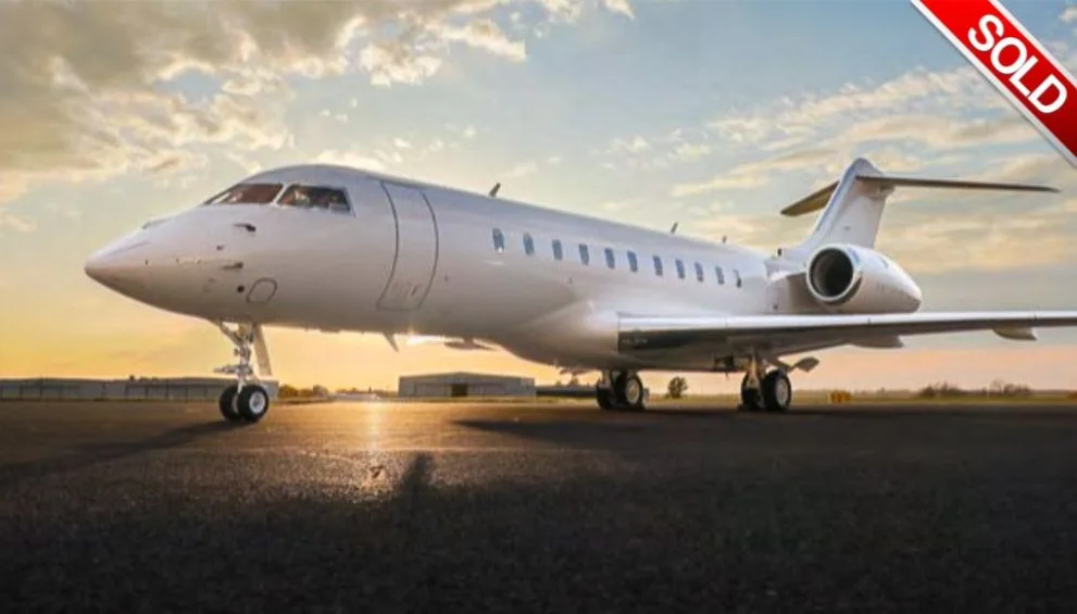 A sleek white business jet, likely a Bombardier Global, is parked on an airfield at sunset.