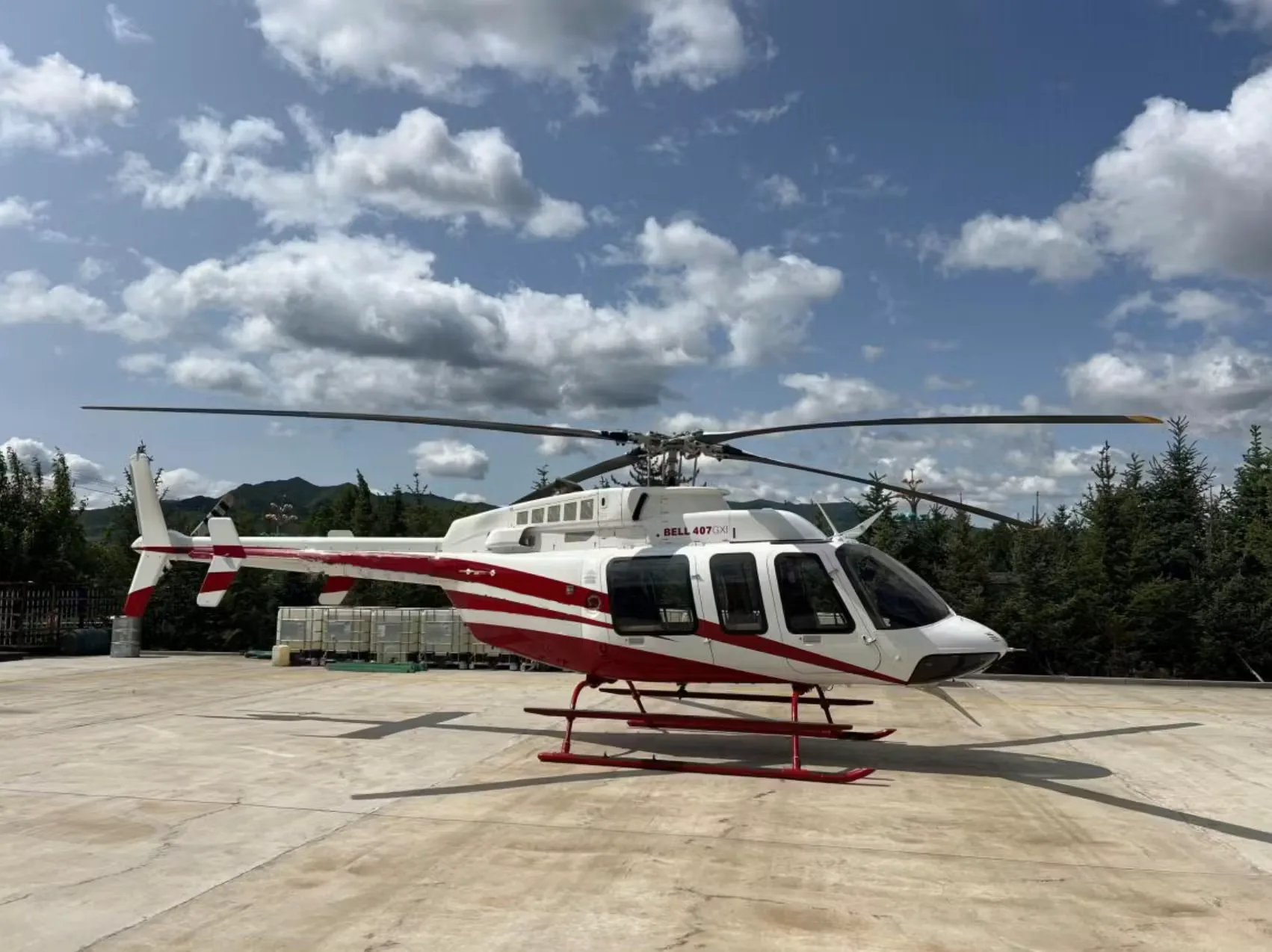 A white and red Bell 407 GXi helicopter parked on a concrete pad surrounded by trees under a cloudy sky.