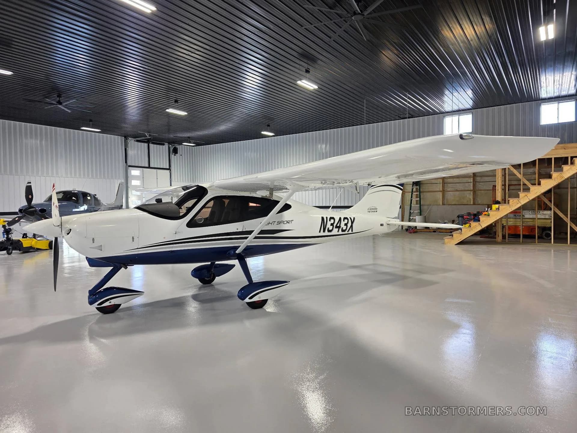 A front-side view of a white Tecnam P2008 Light Sport aircraft with blue accents, registration N343X, parked in a hangar with a wooden staircase in the background.