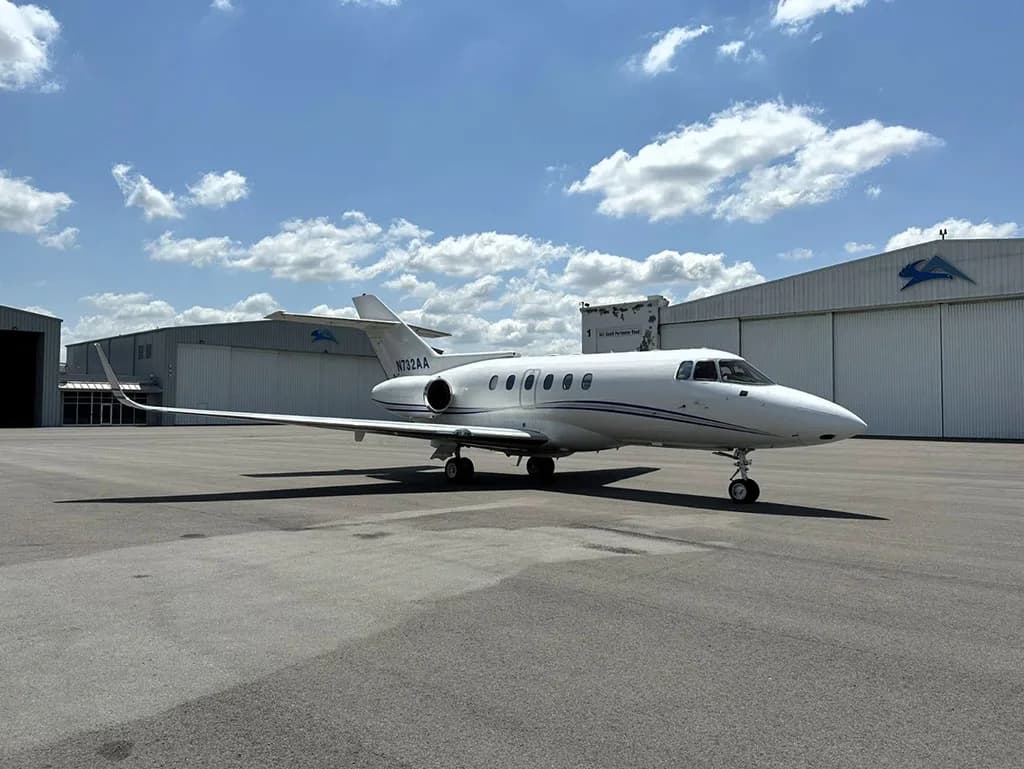 A white business jet, N732AA, with blue stripes parked on an tarmac with hangars in the background under a blue sky with clouds.