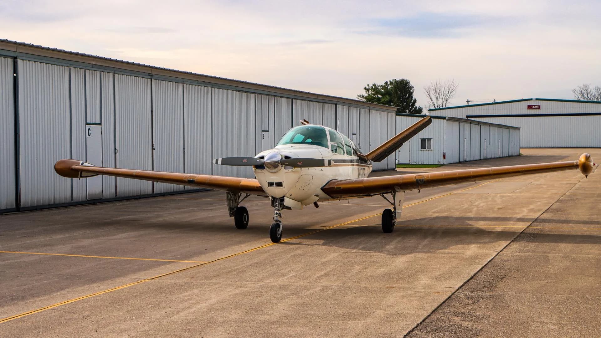 Front three-quarter view of a white and gold Beechcraft Bonanza parked on an airfield.