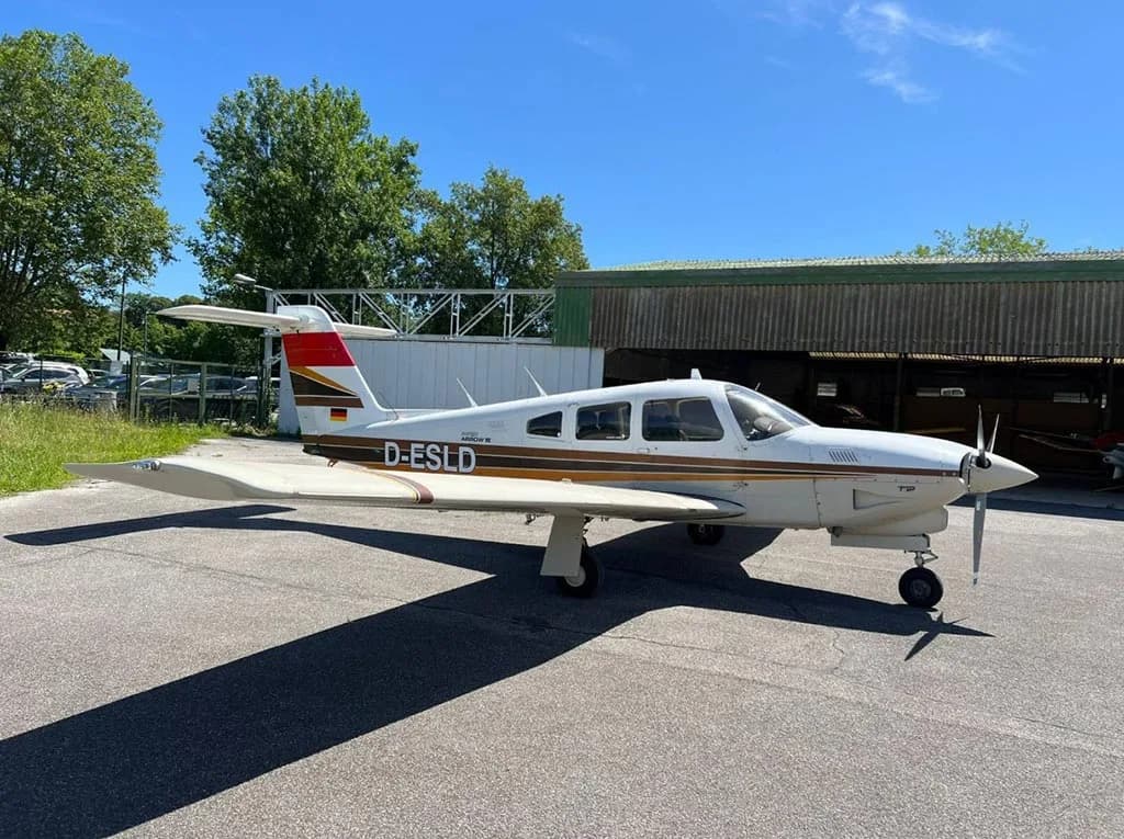 A white and brown Piper Arrow III (D-ESLD) parked on an apron next to a green hangar.