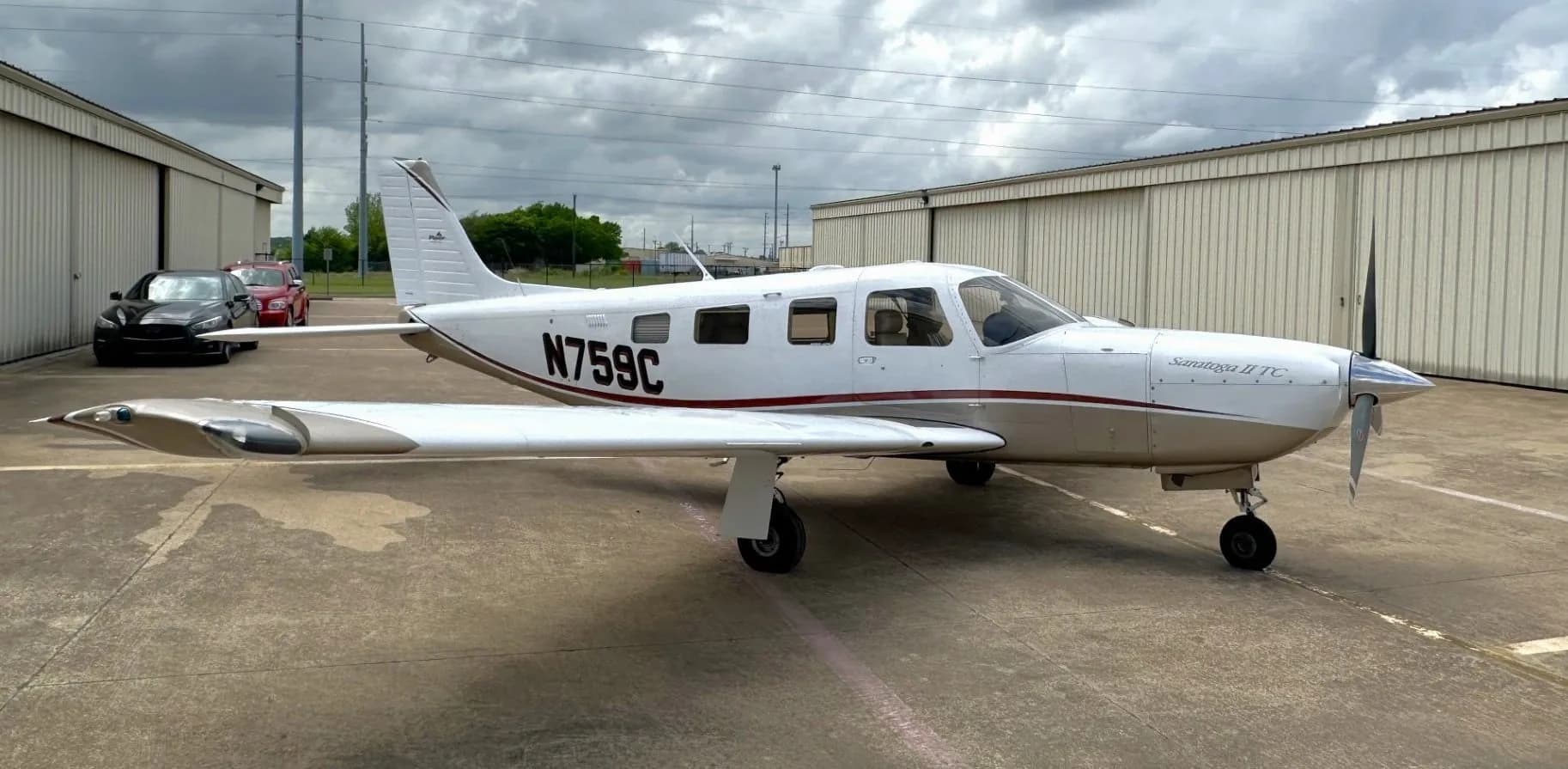 A white and tan Piper Saratoga II TC aircraft (N759C) parked on an asphalt ramp in front of hangars under a cloudy sky.