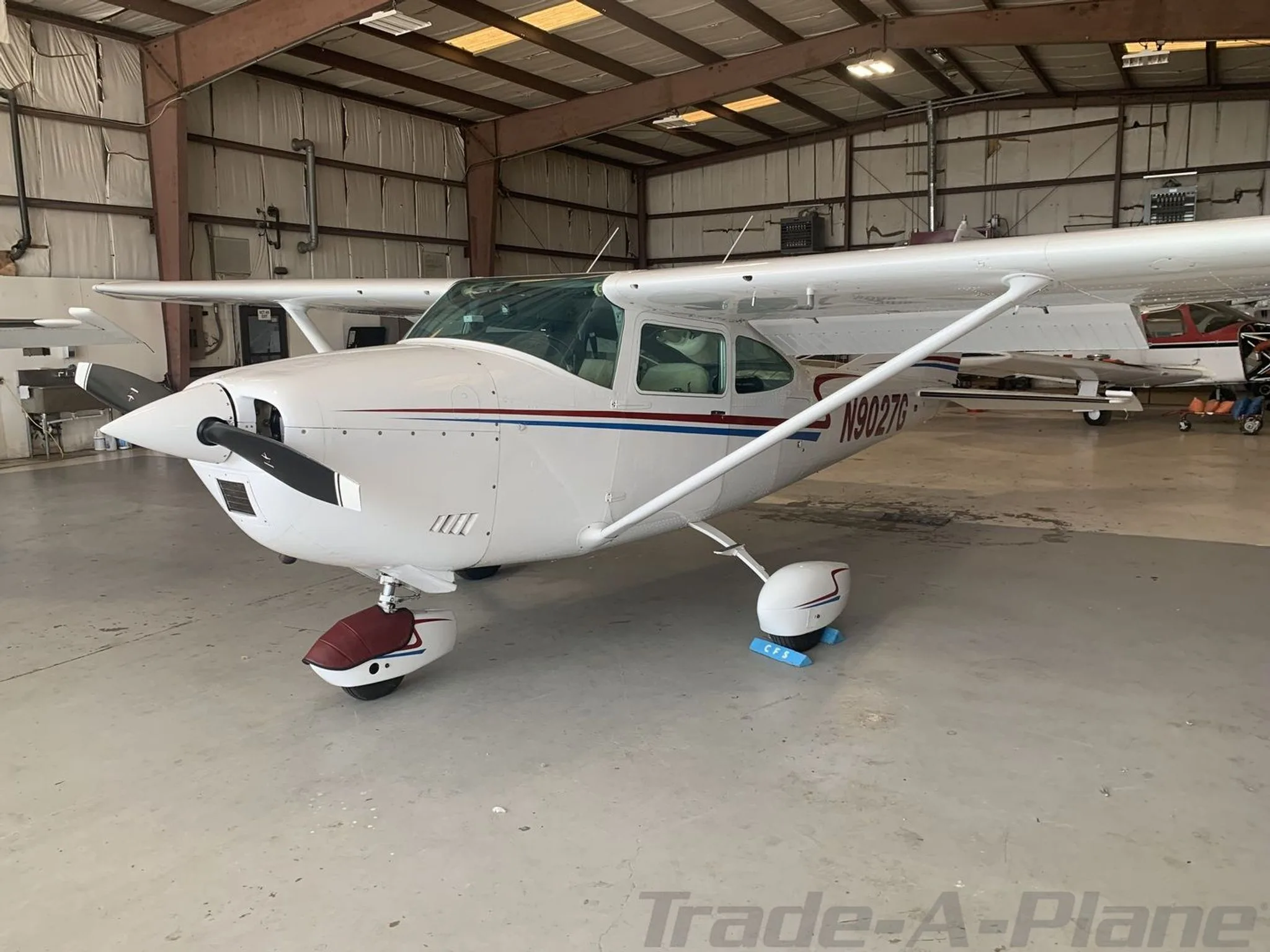 A white Cessna 182N Skylane with red and blue stripes, registration N9027G, parked inside a metal hangar.