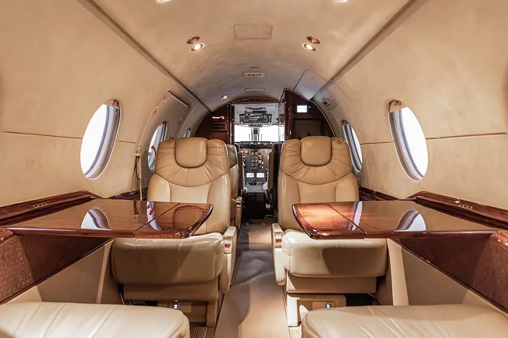 View down the aisle of a private jet cabin with light-colored leather seats and polished wood tables extended.