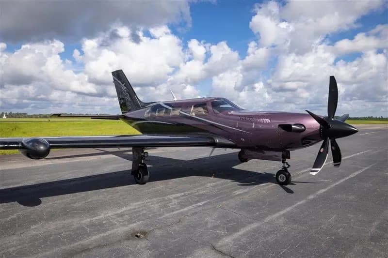 A purple and black Piper M600 turboprop aircraft is parked on a tarmac under a partly cloudy sky.