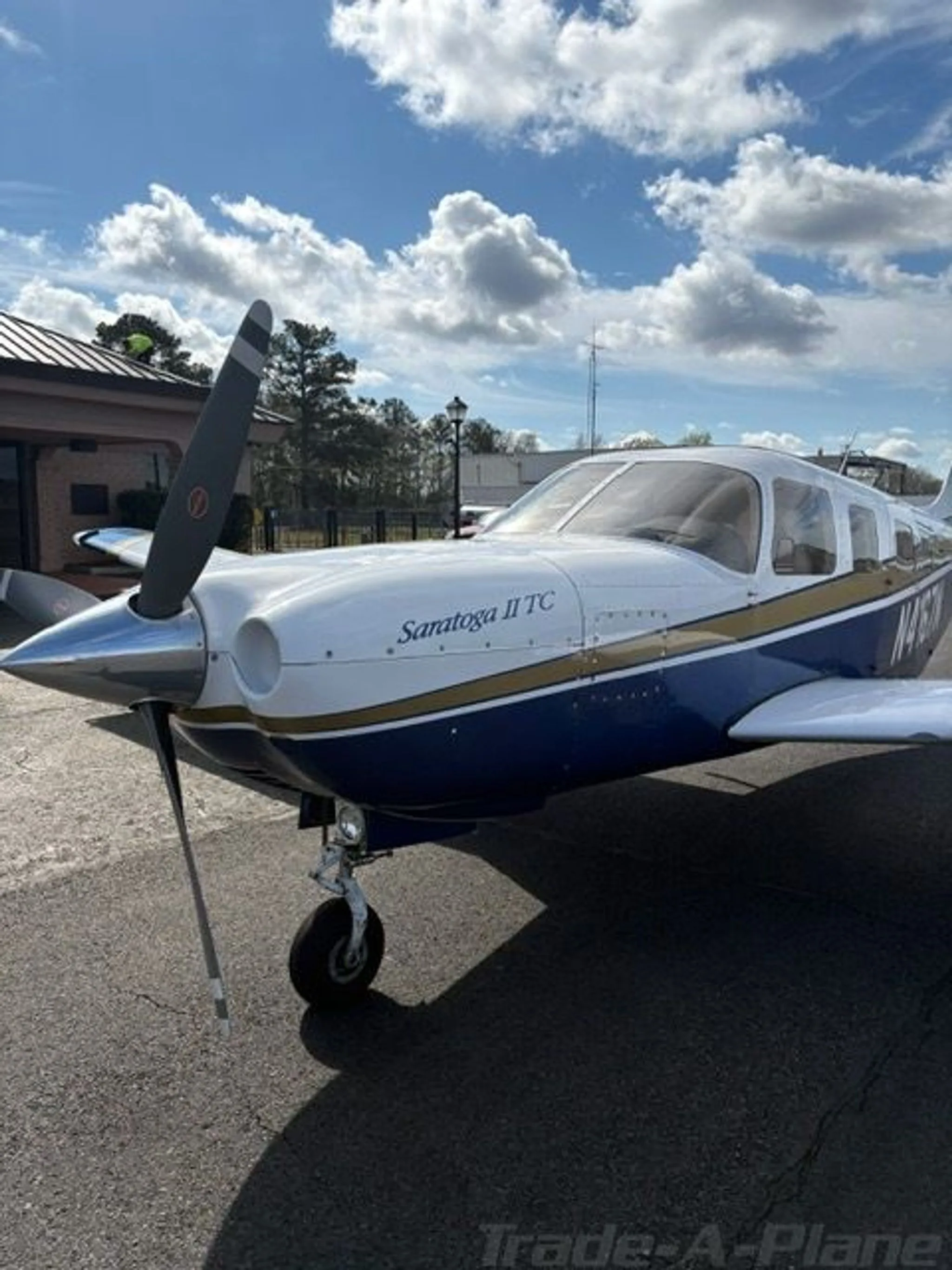 A front three-quarter view of the Piper Saratoga II TC aircraft, N4167N, with its propeller and white, blue, and gold paint, parked near a building.