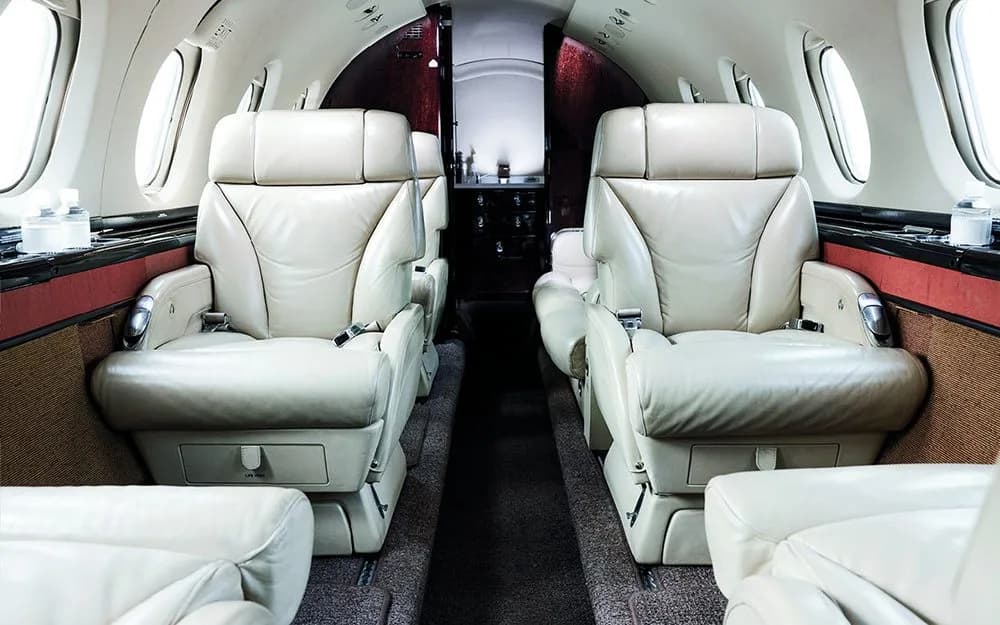 The aircraft cabin shows two rows of cream-colored leather seats with oval windows and a view towards the rear.