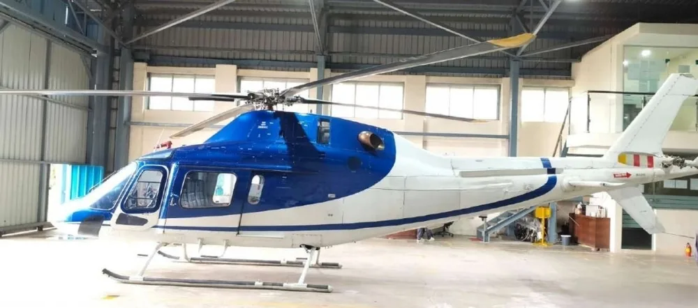 Side view of a blue and white helicopter with its main rotor blades extended, parked inside a hangar.
