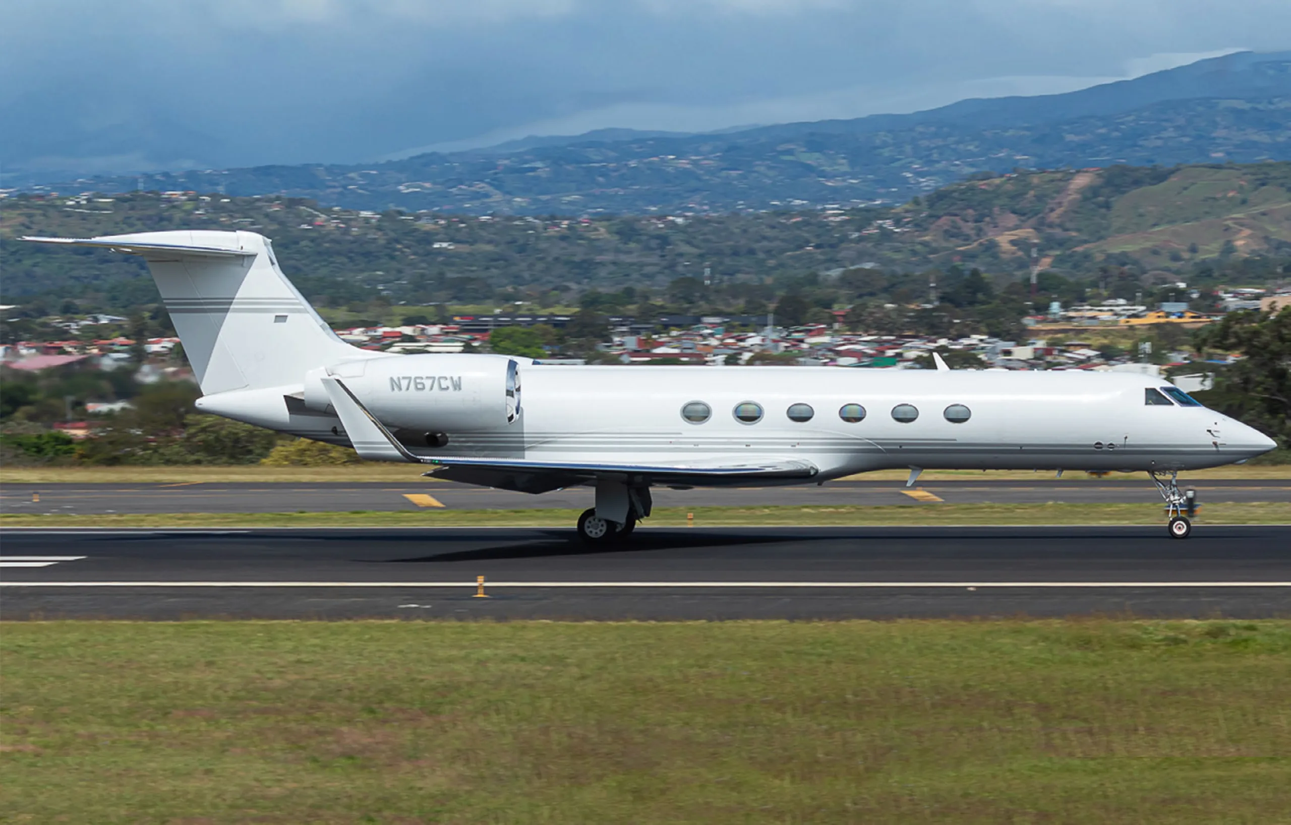 A white Gulfstream G550 business jet with registration N767CW taxiing on a runway with mountains in the background.