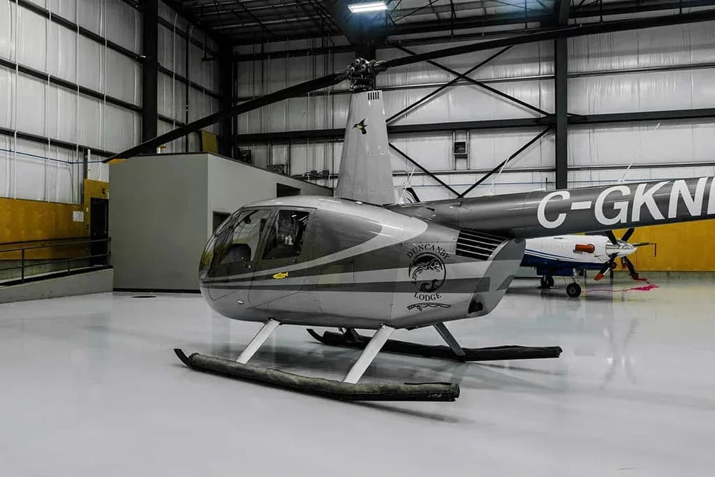 A grey and white Robinson R44 helicopter with "DUNCANBY LODGE" logo, seen from the front-right in a hangar.