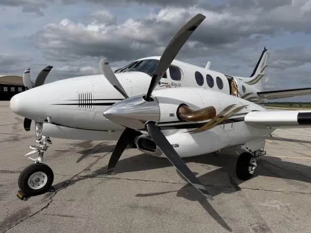 A white Beechcraft King Air twin-turboprop aircraft with gold and black stripes parked on an asphalt tarmac under a cloudy sky.