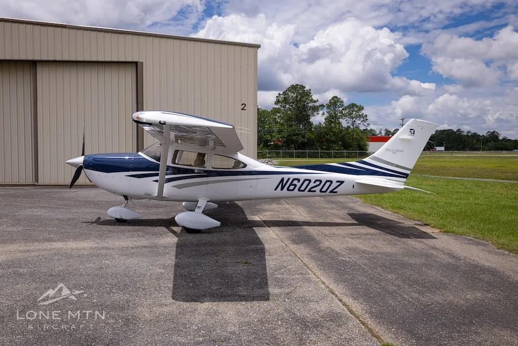 A right-side view of a white and blue Cessna Skylane TC (N6020Z) parked on an airfield next to a hangar.