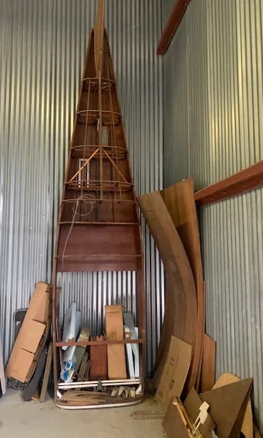 The wooden skeletal frame of an aircraft fuselage or large component stored against a corrugated metal wall.