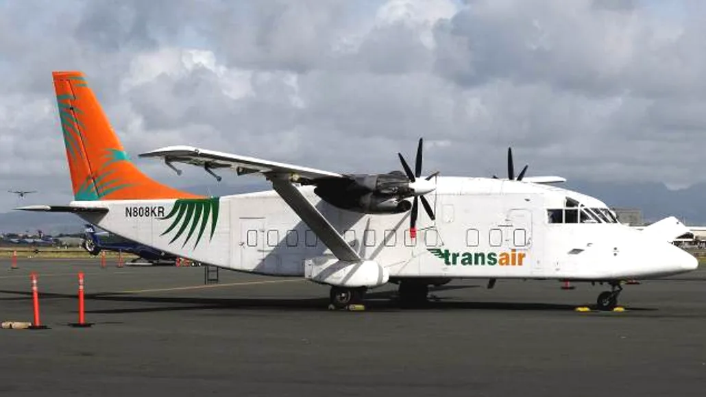 A Transair aircraft parked on the tarmac with mountains in the background.
