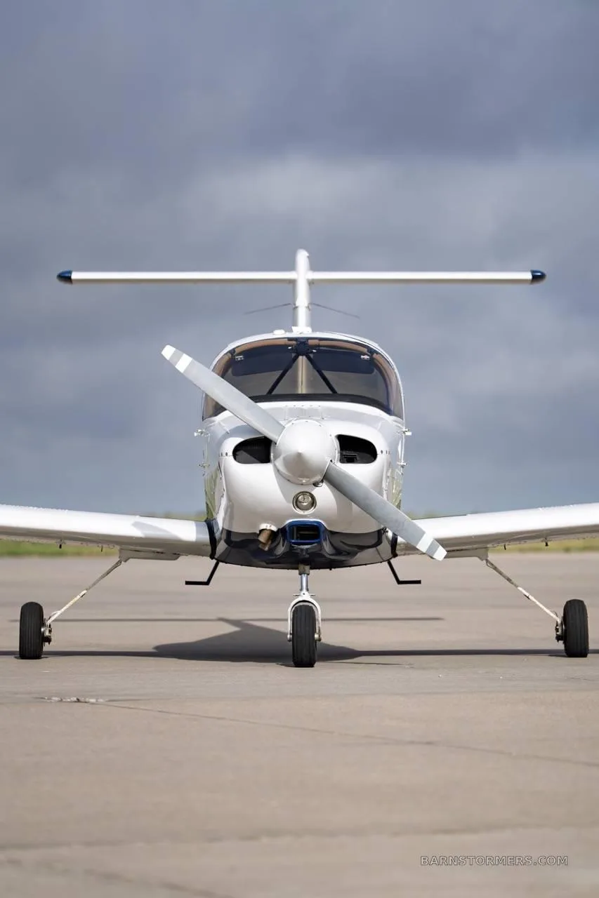 A white and blue single-engine propeller aircraft is parked on an asphalt runway, viewed directly from the front under a cloudy sky.