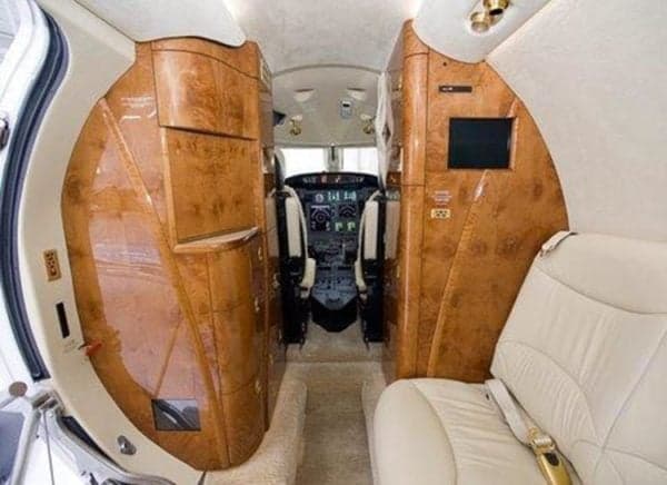 The luxurious interior of a private jet cabin features light leather seats, polished wood cabinetry, and a view into the cockpit.