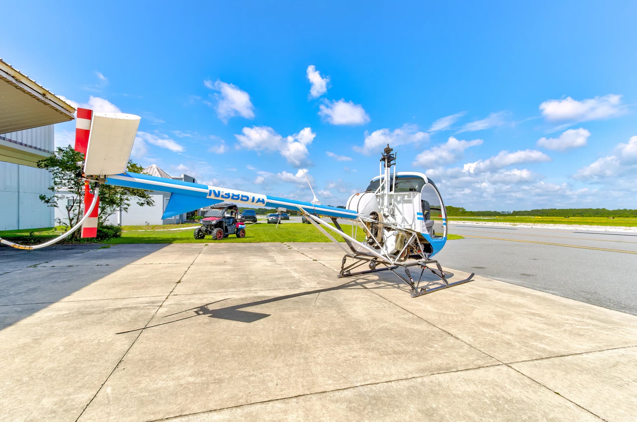 A blue and white Schweizer 300CBi helicopter, N385TA, parked on an airfield tarmac with a hangar and trees in the background.