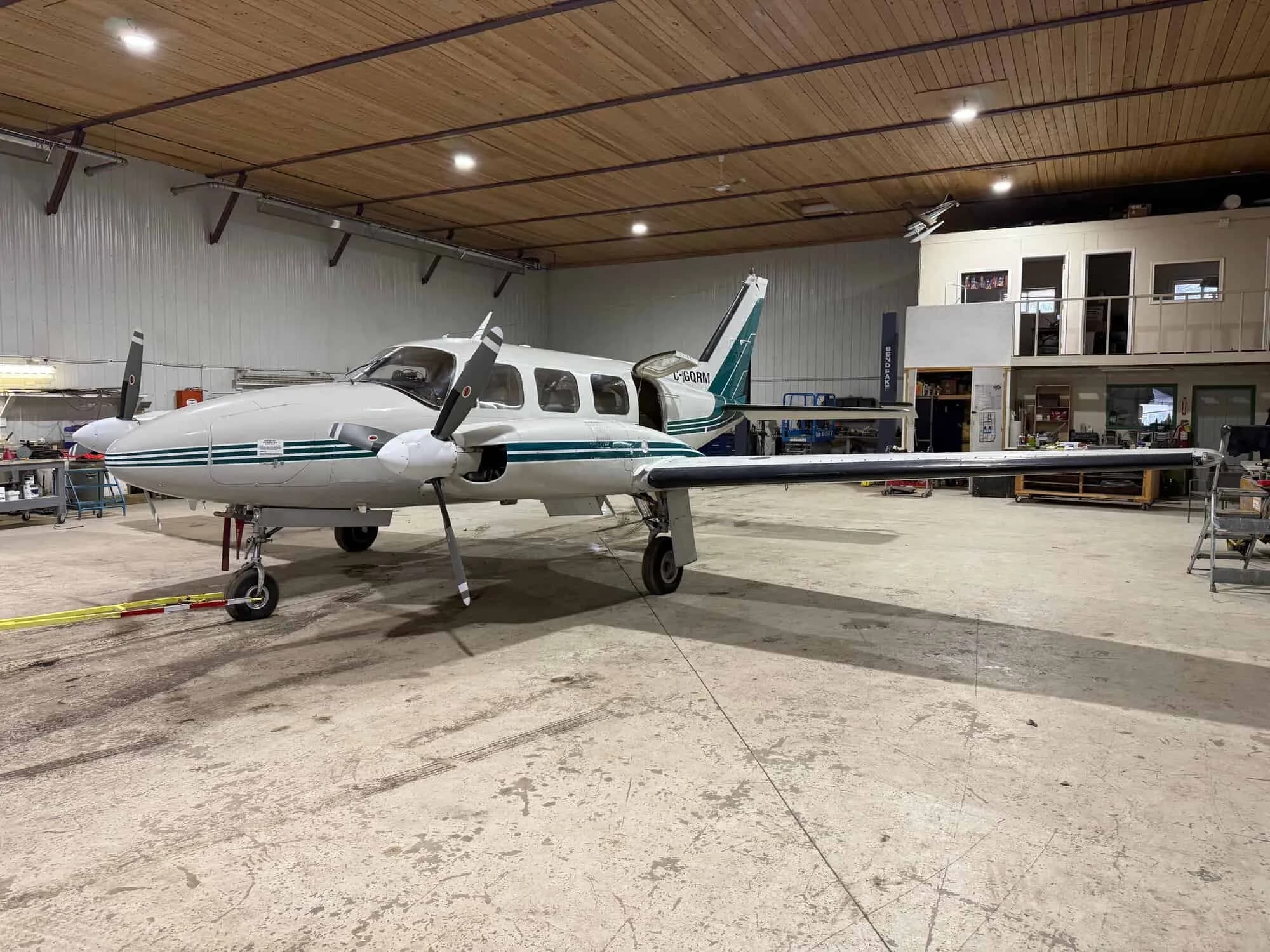 A white and green twin-engine aircraft, registration C-GQRM, parked in a hangar with its nose facing right.