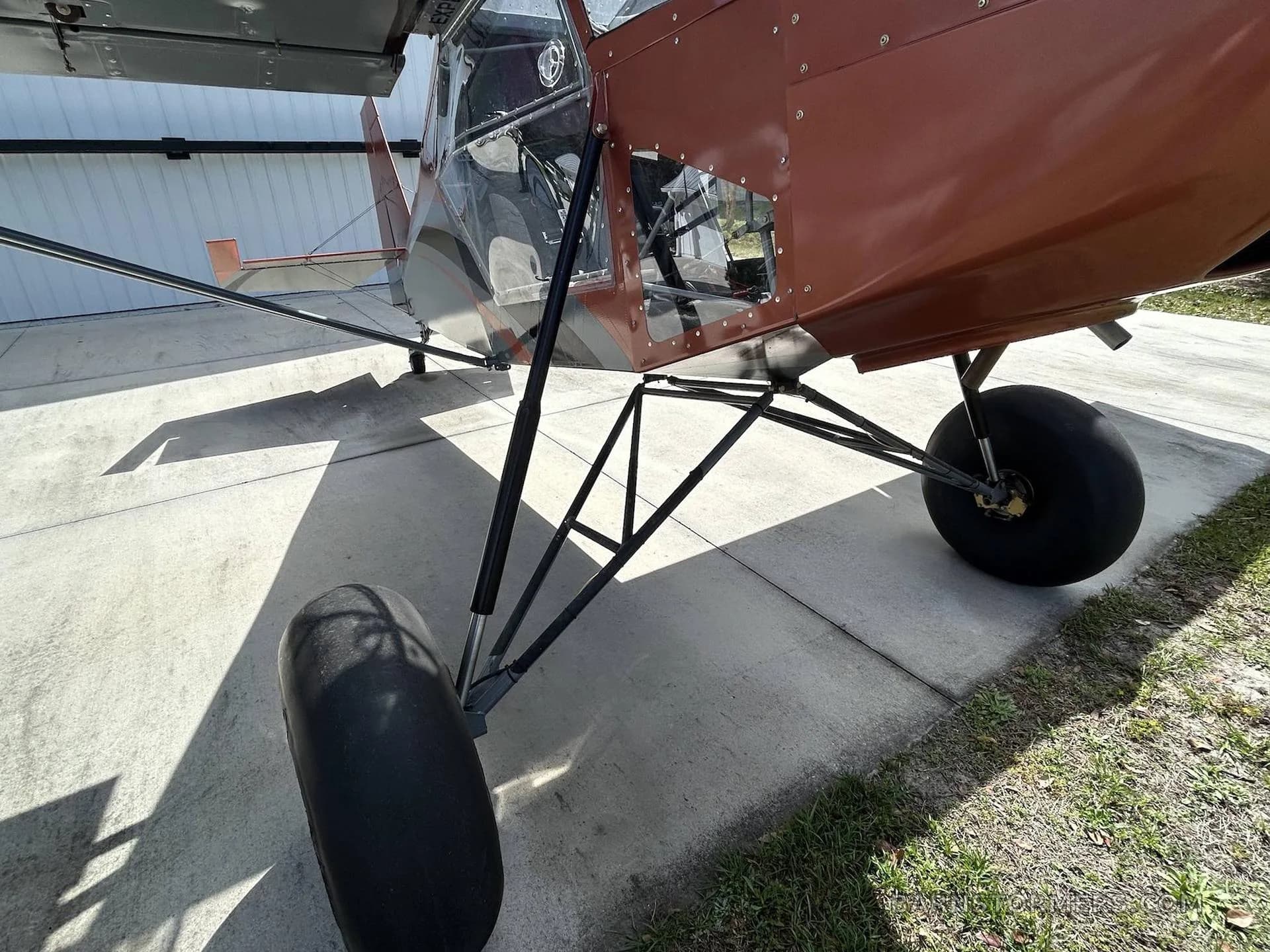 A close-up side view of the orange and grey bush plane, showing its large tires and landing gear on concrete.
