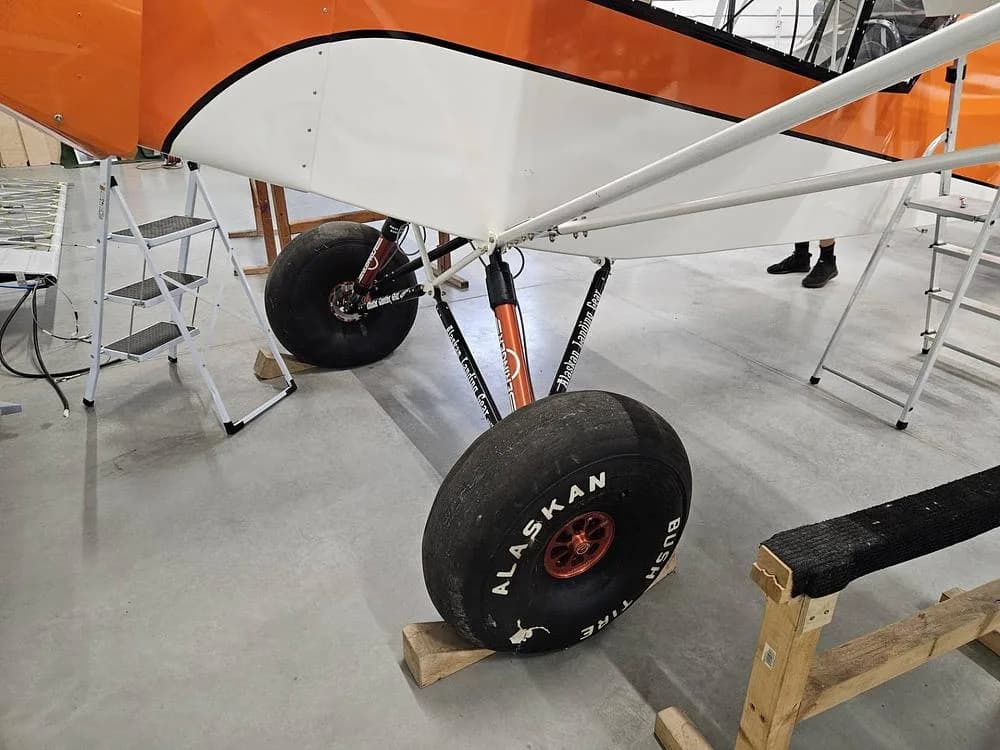 A close-up of the large "Alaskan Bushwheel" tires and landing gear of the orange and white aircraft in a hangar.