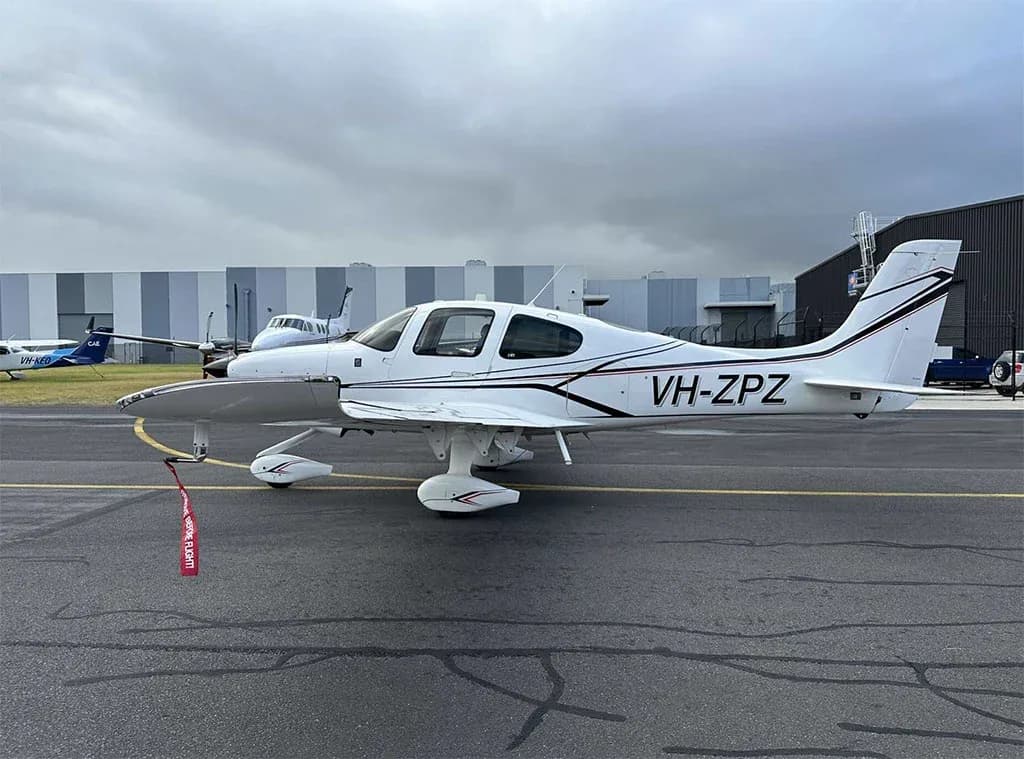 A white Cirrus SR22 aircraft with black and red stripes, registration VH-ZPZ, parked on a tarmac with hangars in the background.