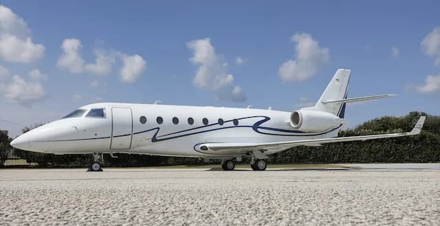 A white business jet with a blue stripe livery is parked on a tarmac under a blue sky with clouds.