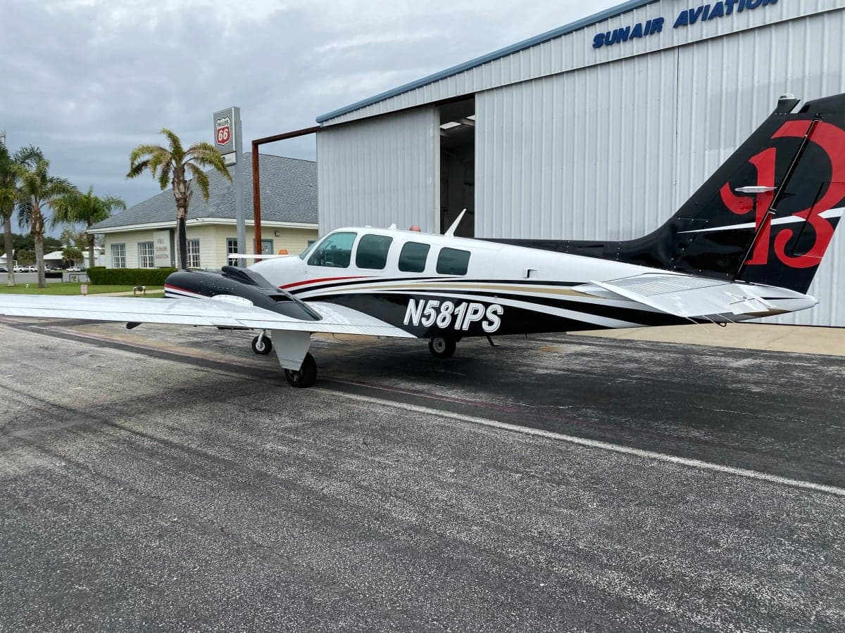 A white and black twin-engine aircraft with gold and red stripes, registration N581PS, parked on an airfield next to a hangar.