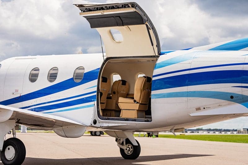 A Pilatus PC-12 aircraft with a blue and white paint scheme is parked on the tarmac with the cargo door open, revealing the interior.