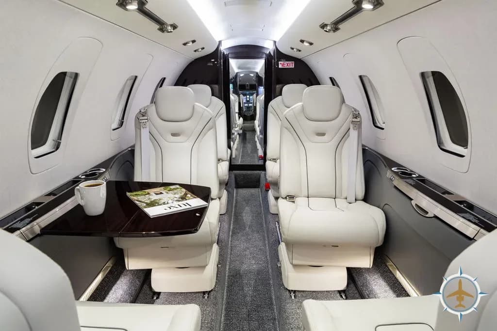 The aircraft cabin interior with two rows of white leather seats facing each other, a table with a magazine and coffee cup.
