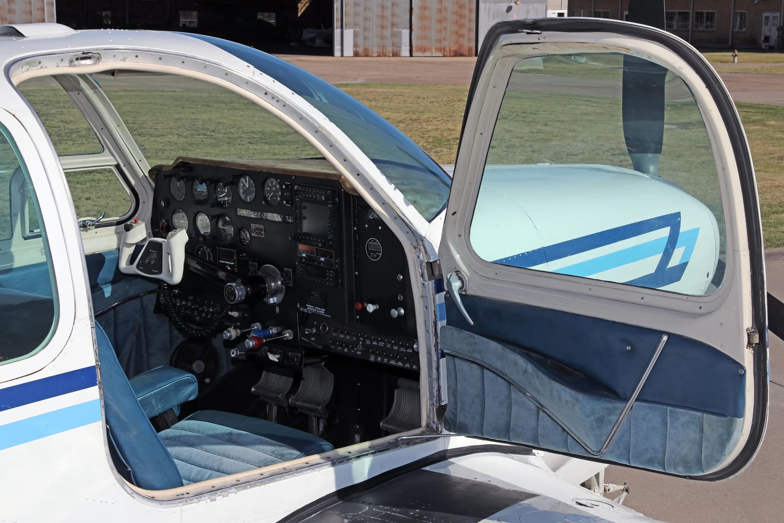 The cockpit of a white aircraft with blue interior, showing the instrument panel and controls through an open door.