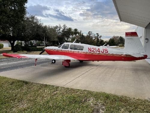 A red and white Mooney M20C aircraft, registration N214JS, parked outside a hangar.