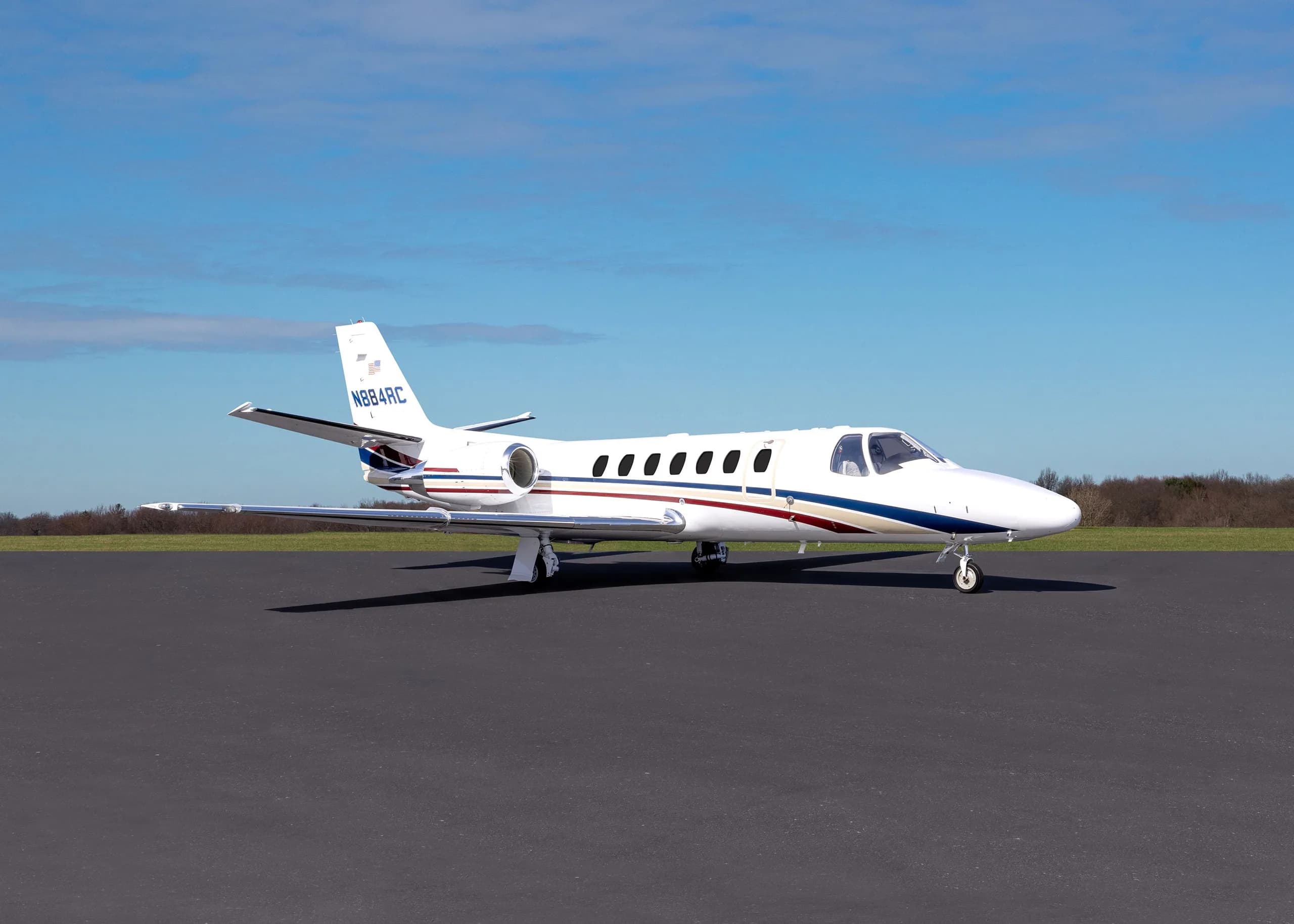 A white business jet with blue and red stripes, registration N884RC, parked on an asphalt tarmac under a blue sky.