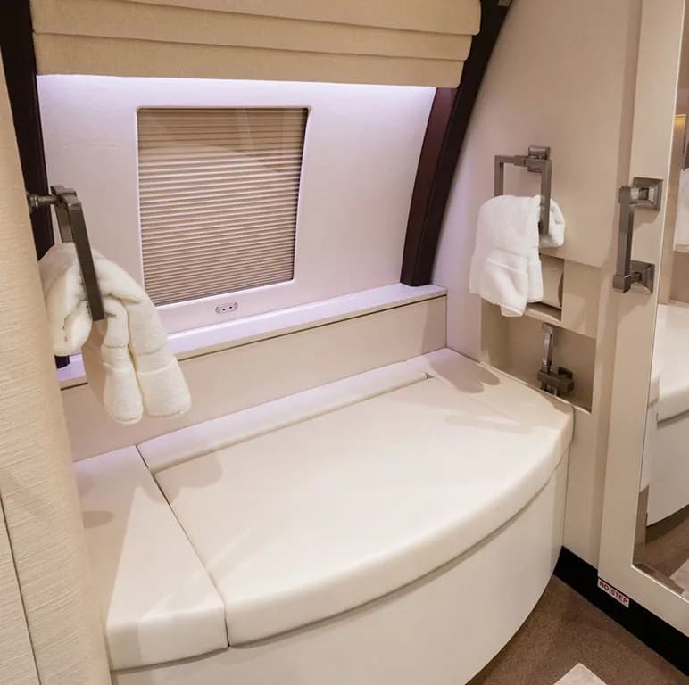 A well-appointed aircraft bathroom with a padded bench seat, a window with blinds, and white towels hanging.