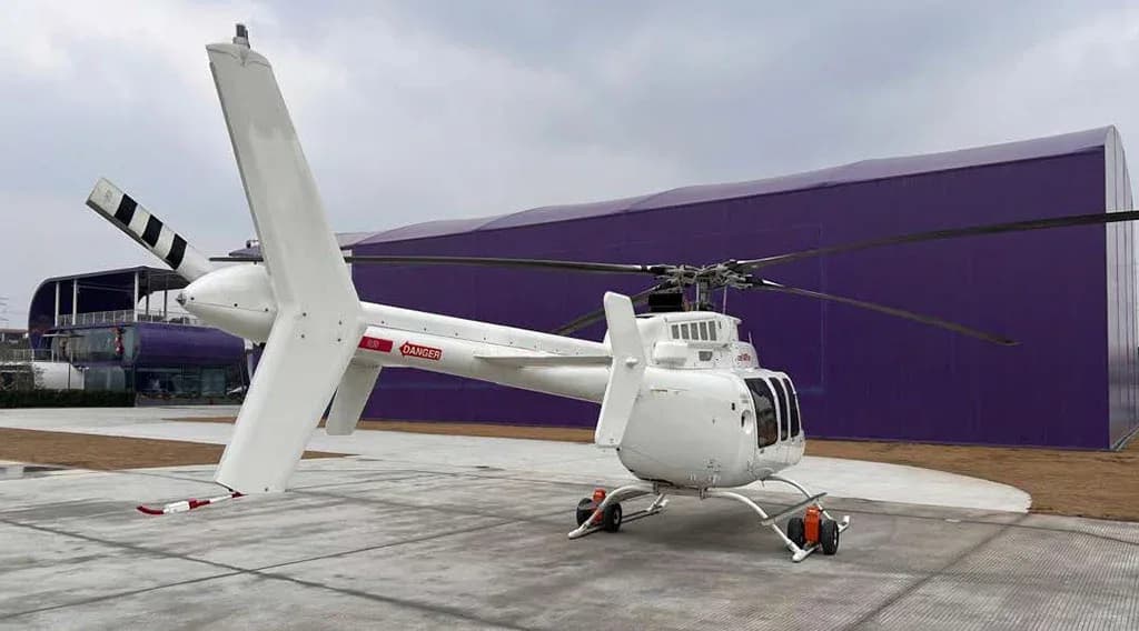A white Bell 407GXP helicopter, viewed from the rear-right, parked on a concrete pad next to a large purple building.