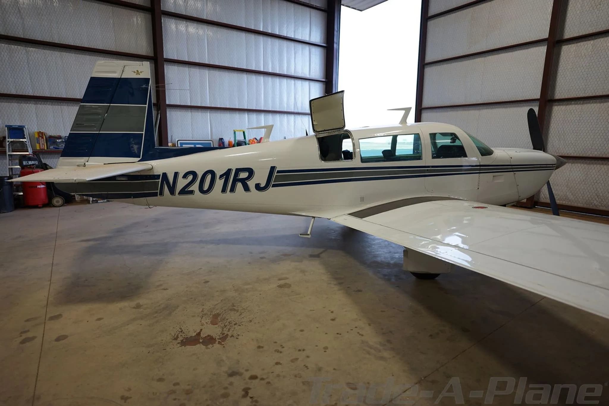 A white single-engine aircraft with blue and grey stripes, registration N201RJ, parked in a hangar with its door open.
