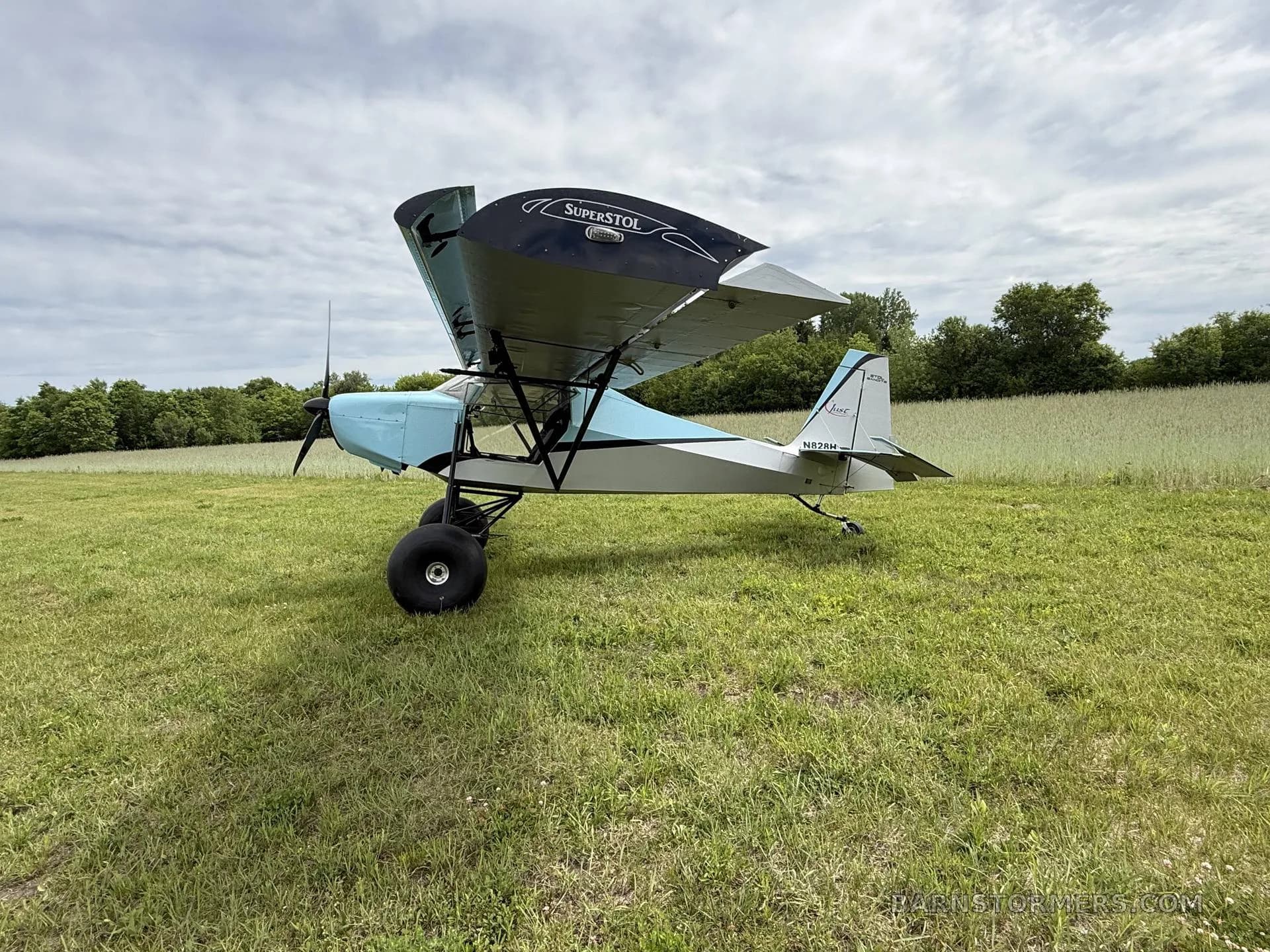 A rear-right view of a light blue and grey Just Aircraft SuperSTOL with its wings folded, parked on a grassy field.