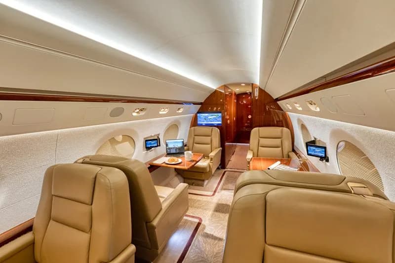 The cabin interior of a business jet with tan leather seats, small tables, and personal entertainment screens.
