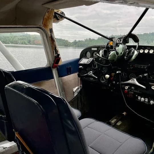 The interior of a floatplane shows the cockpit with instruments and controls, and two grey seats, looking out over water.