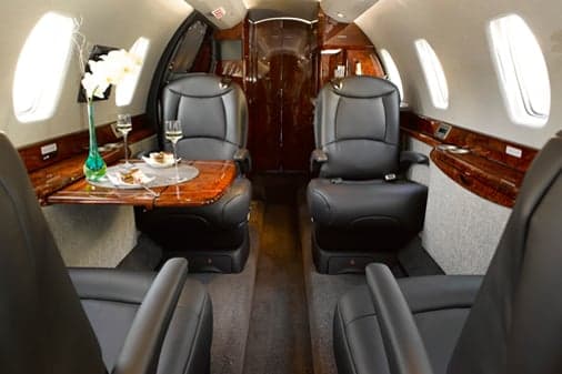 The spacious interior of a private jet cabin with black leather seats, polished wood paneling, and a table adorned with flowers and drinks.