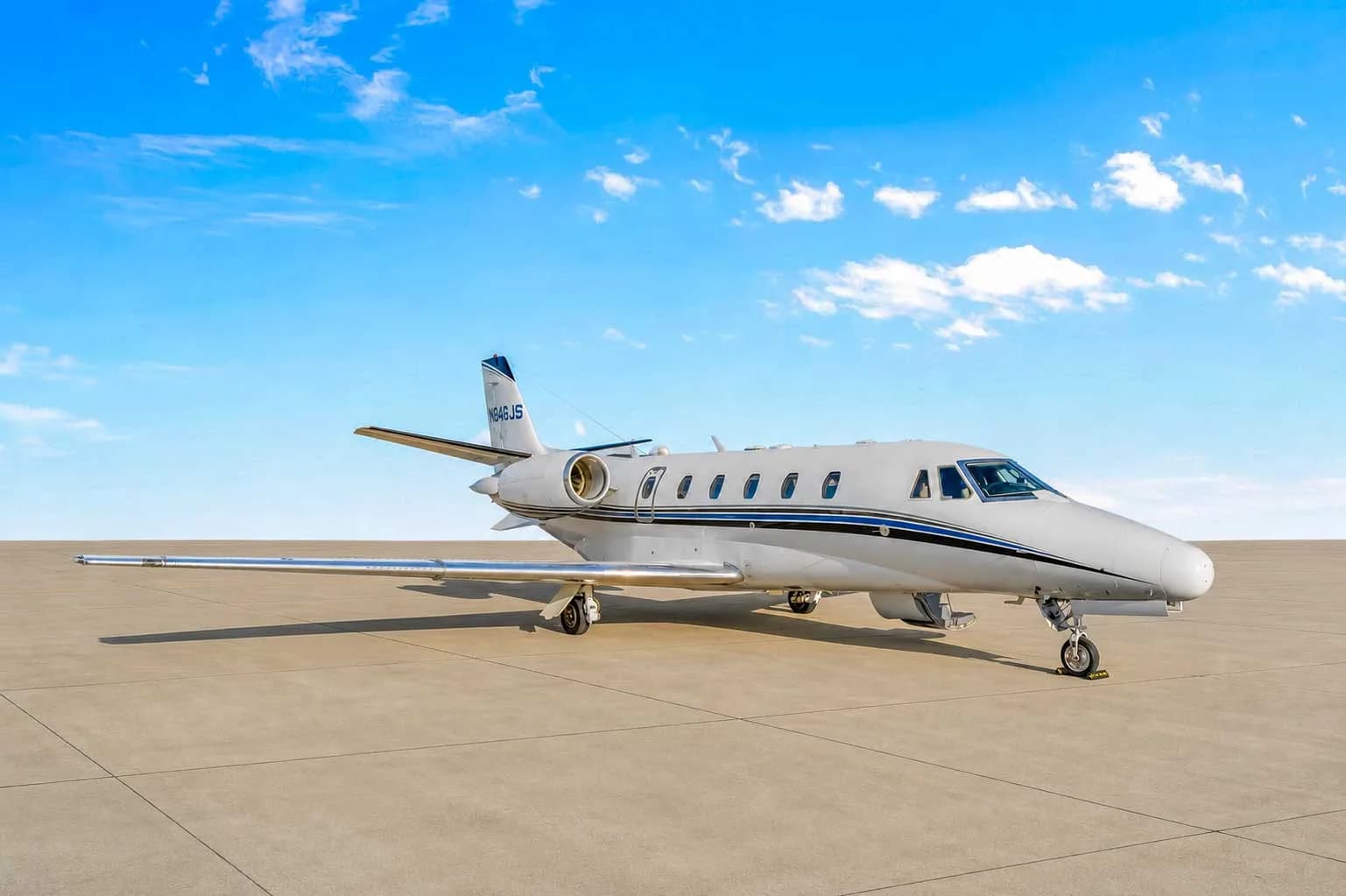 A white Cessna Citation business jet with blue and black stripes, registration N846JS, parked on an airfield under a blue sky.