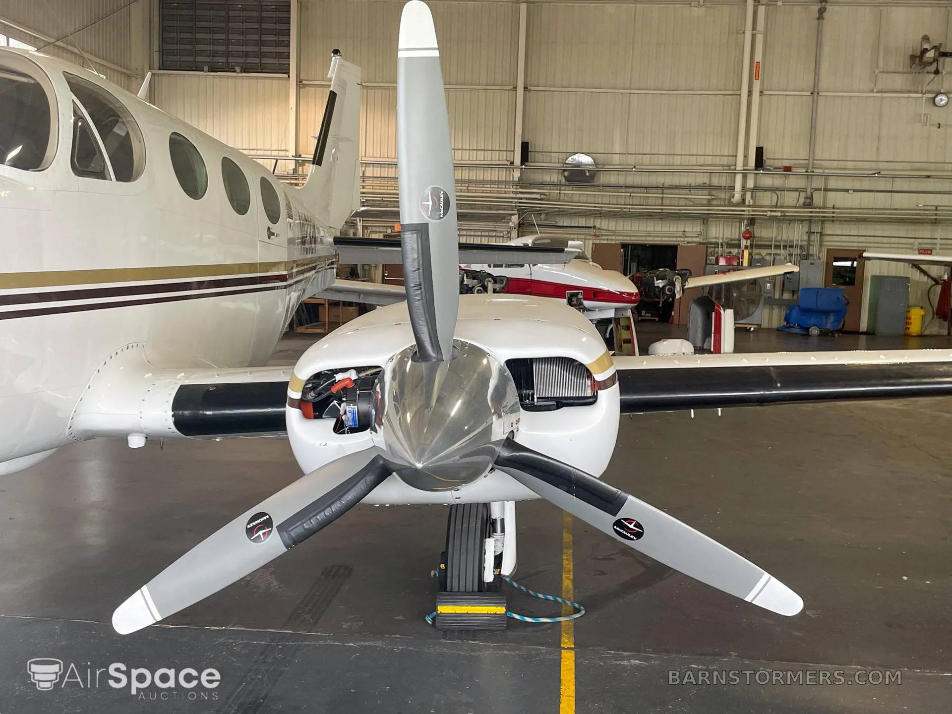 A close-up view of the front propeller and engine cowling of a white twin-engine aircraft inside a hangar.