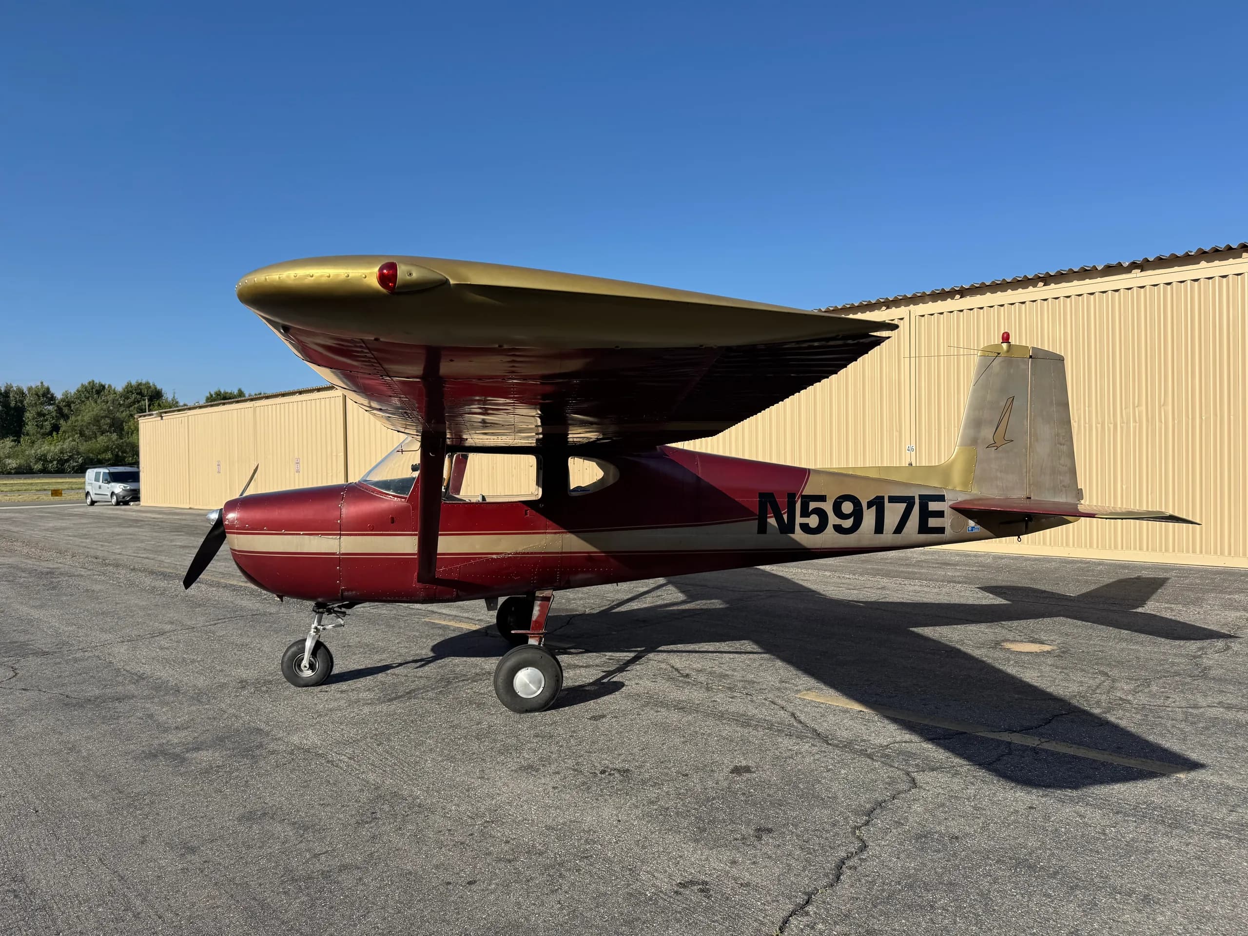 A red and gold vintage high-wing propeller aircraft, N5917E, parked on an asphalt apron next to a hangar under a clear blue sky.