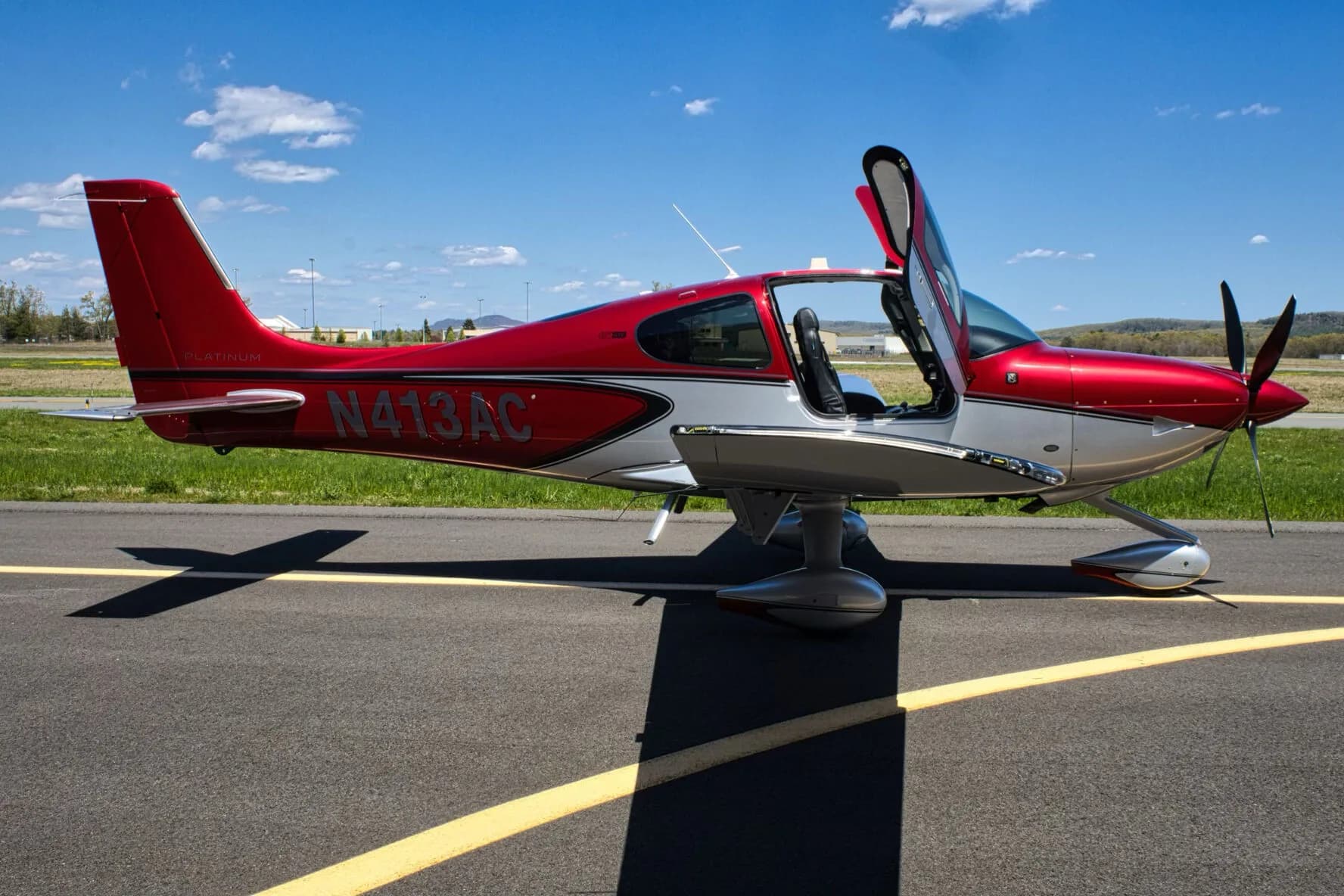 A red and silver Cirrus SR22T aircraft, N413AC, with its gull-wing doors open, parked on an airport tarmac.