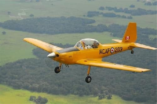 A yellow aircraft with RCAF markings is flying over a green landscape.