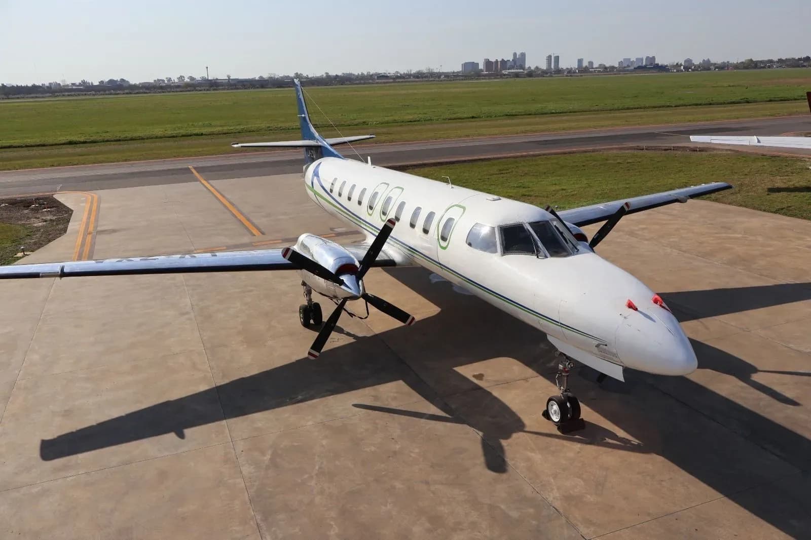 An aerial view of a white turboprop aircraft with blue and green stripes, parked on an tarmac with a city skyline in the background.