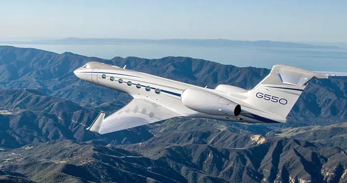 A white Gulfstream G550 business jet with blue accents flying over mountainous terrain and water.