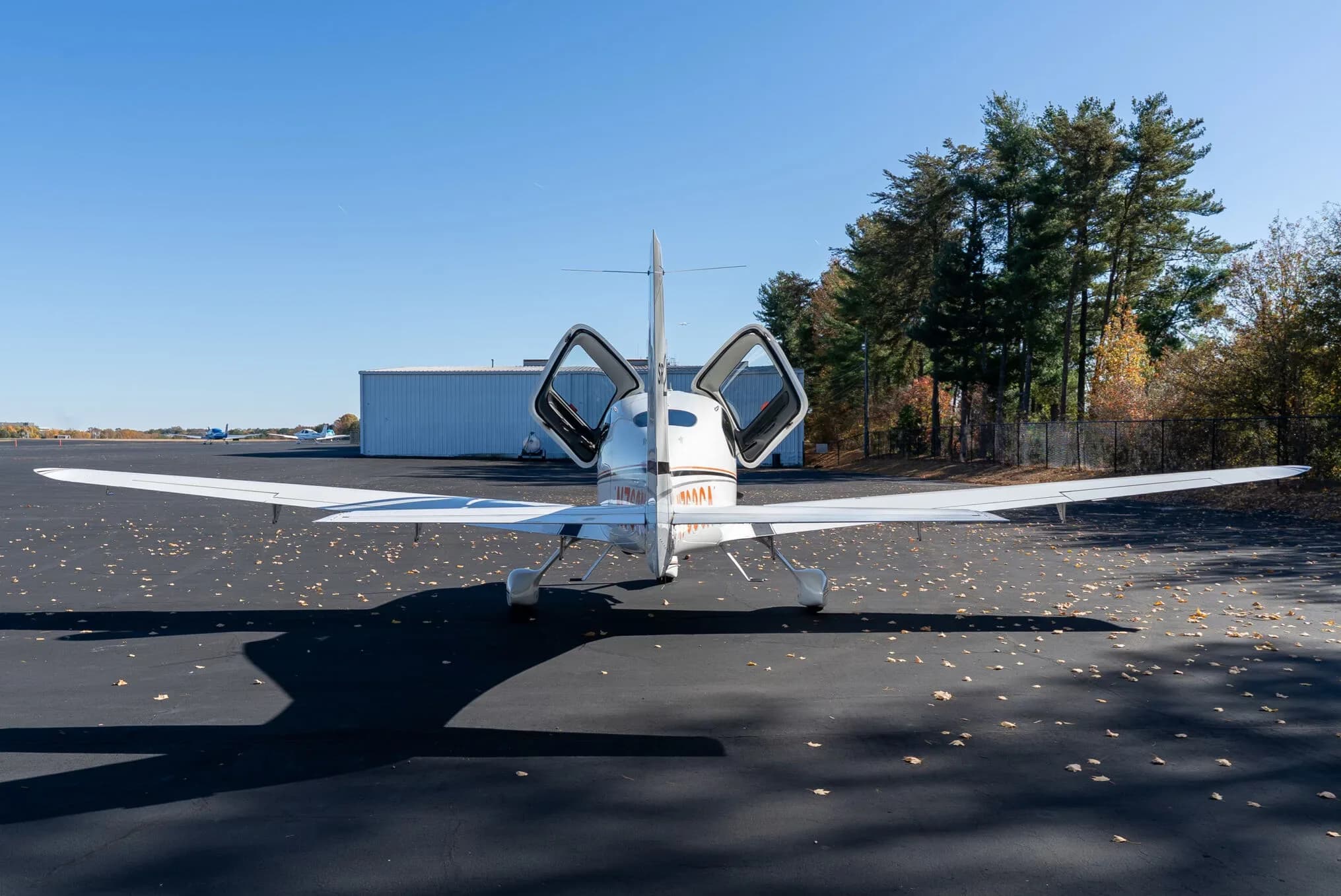 A white Cirrus SR20 aircraft with its doors open is viewed from directly behind, parked on an asphalt tarmac.
