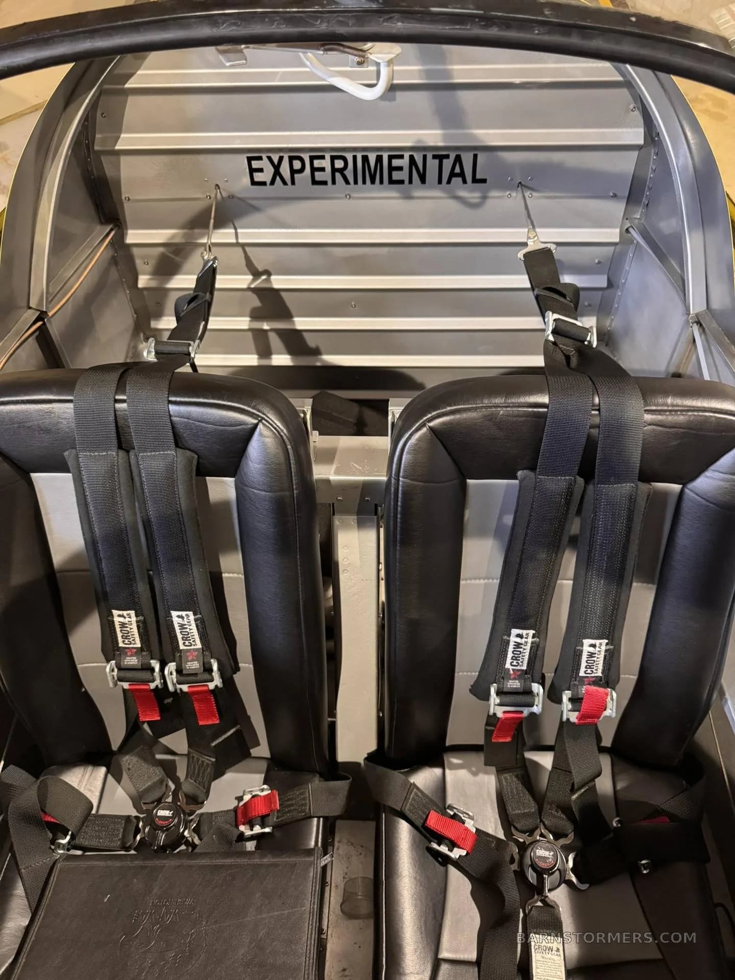 The interior of an experimental aircraft shows two black seats with multi-point harnesses and "EXPERIMENTAL" stenciled on the bulkhead.